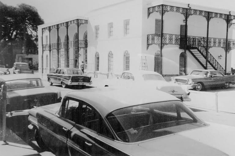 The image is a black and white photograph capturing a scene from the past. It features vintage cars parked on a bustling street in front of a two-story building with ornate balconies.